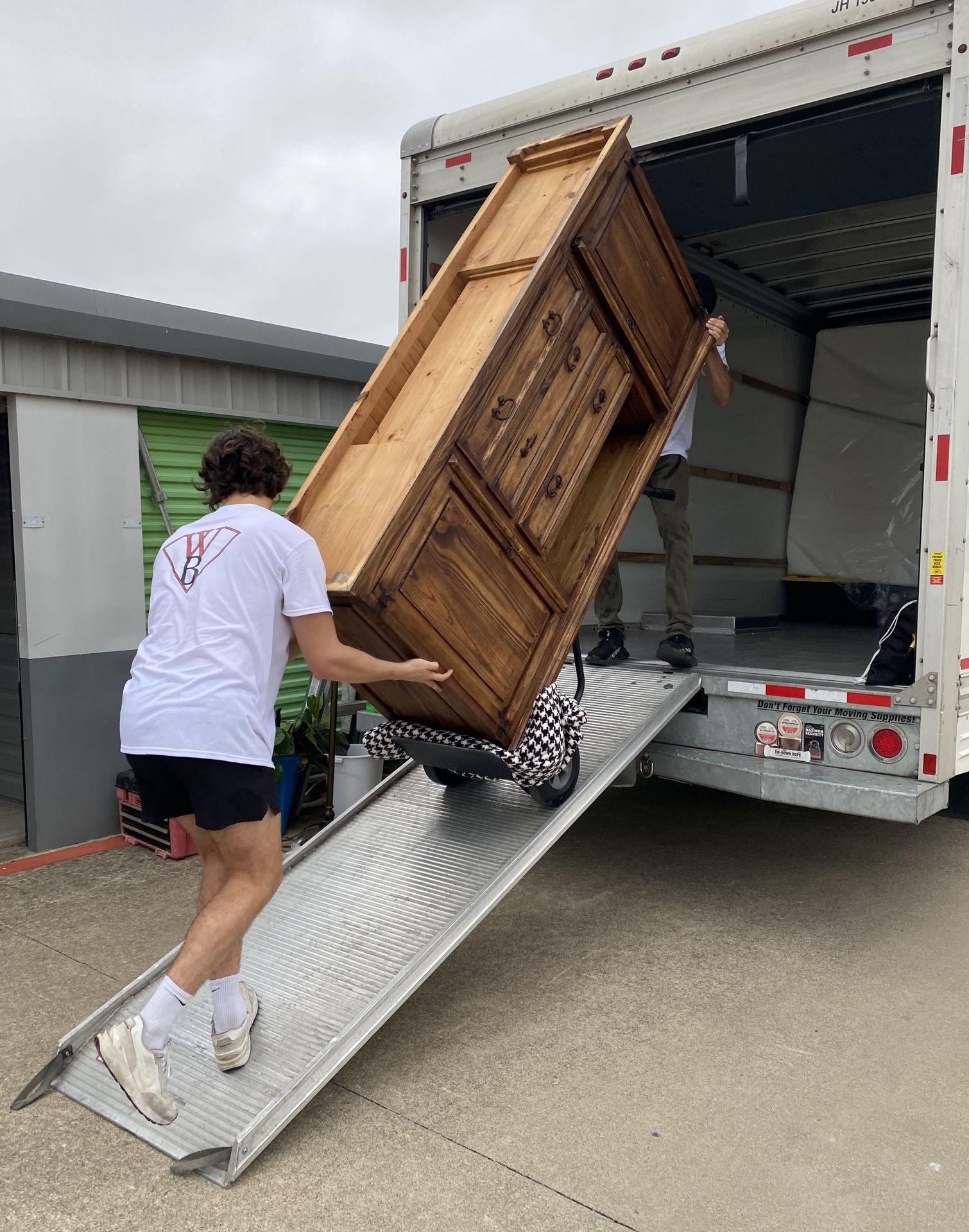 Two people loading a wooden cabinet onto a moving truck using a ramp.